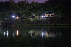 a house is lit up at night by the water's edge with trees in the background