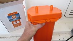 a person holding an orange plastic container in front of a box with the lid open