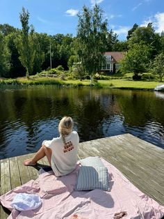 a woman sitting on top of a wooden dock next to a body of water with trees in the background