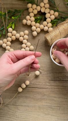 two hands are holding beads and string on a wooden table next to some sprigs