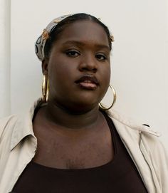 a woman with large hoop earrings and a brown shirt is standing in front of a white wall