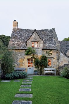 an old stone house is surrounded by greenery and trees in the front yard, with benches on either side