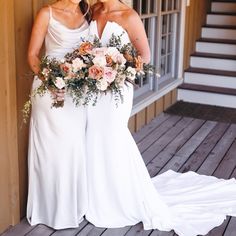 two women in white dresses standing next to each other with flowers on their heads and holding bouquets