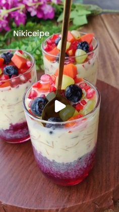 two glasses filled with fruit and yogurt on top of a wooden table next to flowers