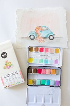 an open watercolor box next to a small painting kit on a white table top