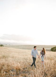 a man and woman holding hands while walking through an open field with tall dry grass