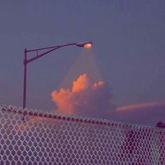 a street light on top of a chain link fence with clouds in the sky behind it