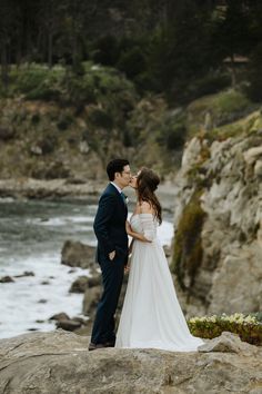 a bride and groom kissing on the rocks by the ocean