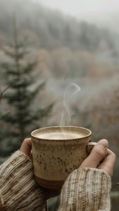 a person holding a coffee cup with steam rising out of it in front of some trees