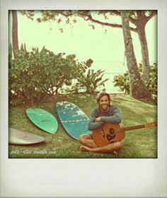 a man sitting in the grass with his guitar and surfboards