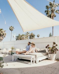 a man and woman sitting on top of a white couch under an awning next to palm trees