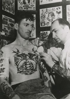 an old photo of two men with tattoos on their chest and arms, looking at each other