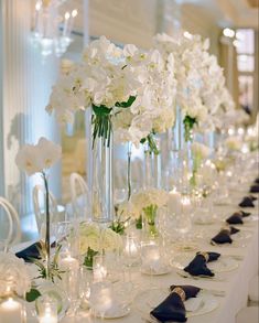 a long table with white flowers in tall vases and candles on each side, along with place settings