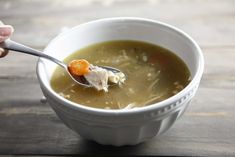 a person holding a spoon full of soup with chicken and carrots in it on a wooden table