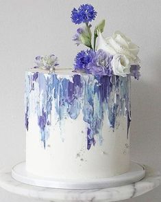 a white cake with blue and purple flowers on top is sitting on a marble stand