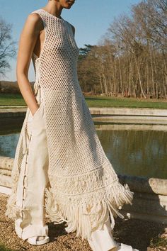 a woman standing in front of a pond wearing a crocheted dress and white shoes