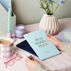 a person holding a book on top of a table next to a vase with flowers