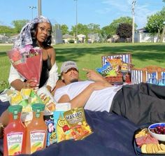 a man and woman laying on a blanket in the grass with food, drinks and snacks