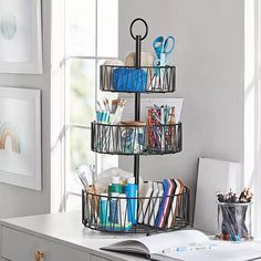 three tiered wire basket with pens, pencils and markers on it in front of a window