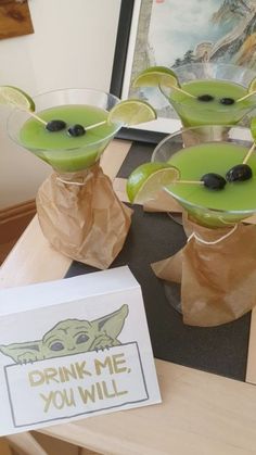 three green drink glasses with olives in them sitting on a table next to a sign that says drink me you will