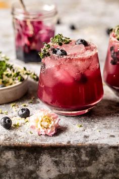 two glasses filled with blueberries and ice on top of a stone table next to flowers