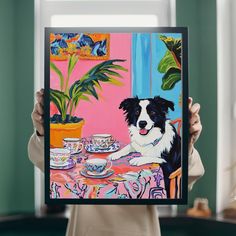 a woman holding up a painting of a dog in front of a table with tea cups and saucers