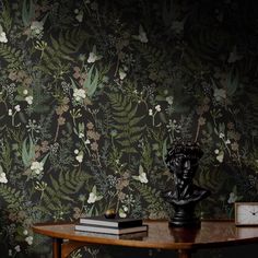 a wooden table topped with a clock next to a wallpaper covered in leaves and flowers