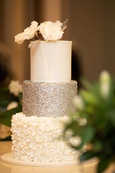 a three tiered white and silver wedding cake with flowers on top, sitting on a table