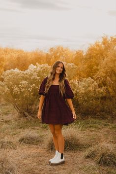 a woman standing in the middle of a field with tall grass and bushes behind her