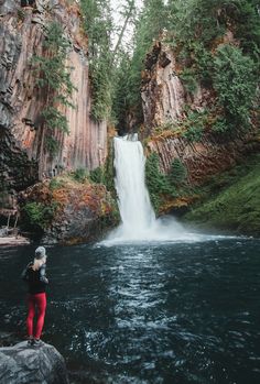 a woman standing on top of a rock next to a river with a waterfall in the background