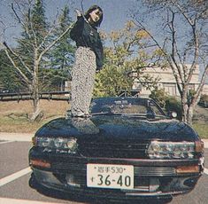 a person standing on the hood of a car in a parking lot with trees behind them