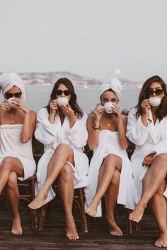 three women in white robes sitting next to each other on a wooden deck near the water