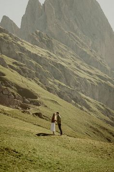 a man and woman standing on top of a lush green field next to a mountain