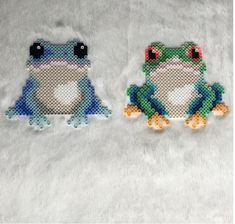 two cross stitch frogs sitting next to each other on a white surface with one frog looking at the camera