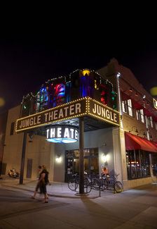 people walking on the sidewalk in front of a theater building at night with colorful lights