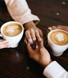 two people holding hands while sitting at a table with cups of coffee in front of them