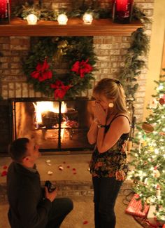 a man kneeling down next to a woman in front of a fire place with christmas decorations