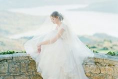 a woman in a wedding dress is sitting on a stone wall with her veil over her head