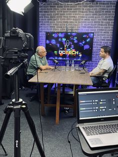 two men sitting at a table in front of a laptop computer and camera set up