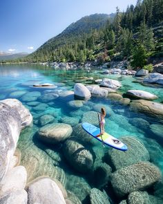 a woman is standing on a surfboard in the clear water near rocks and trees