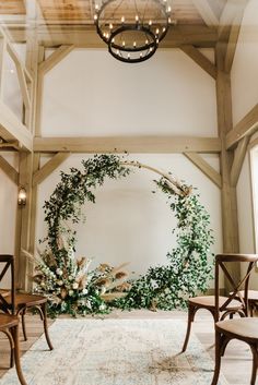 an indoor wedding ceremony with greenery on the wall and wooden chairs in front of it