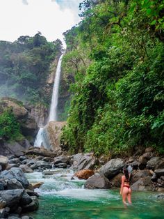 a woman standing in the water next to a waterfall with green trees and rocks on either side