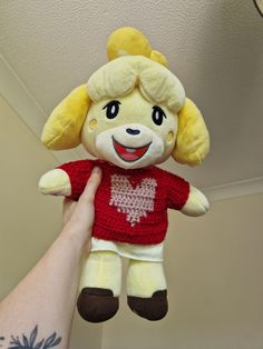 a hand holding a yellow stuffed animal with a heart on it's chest and wearing a red shirt