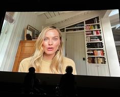 a woman is on the television screen in front of some bookshelves and she has long blonde hair