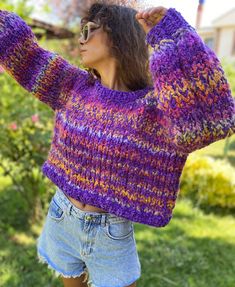 a woman wearing a purple sweater and denim shorts with her hands on her head while standing in the grass