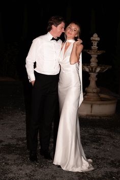 a man and woman in formal wear standing next to each other at night with their arms around each other