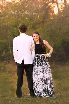 a young man and woman standing next to each other in the grass with trees behind them