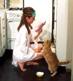 a woman kneeling down next to a cat on the floor with a bowl in front of her