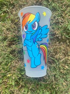 a plastic cup sitting in the grass with a rainbow pony on it's side