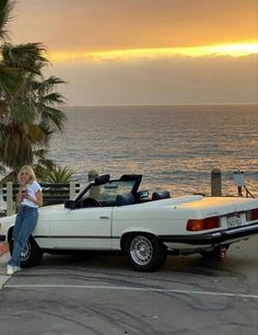 a woman leaning on the hood of a white convertible car near the ocean at sunset